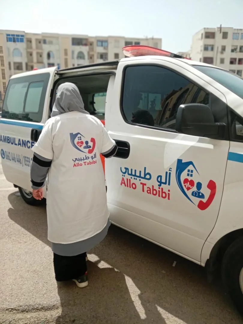 عاملة من ألو طبيبي بمئزر أبيض يحمل الشعار بجانب سيارة إسعاف الشركة